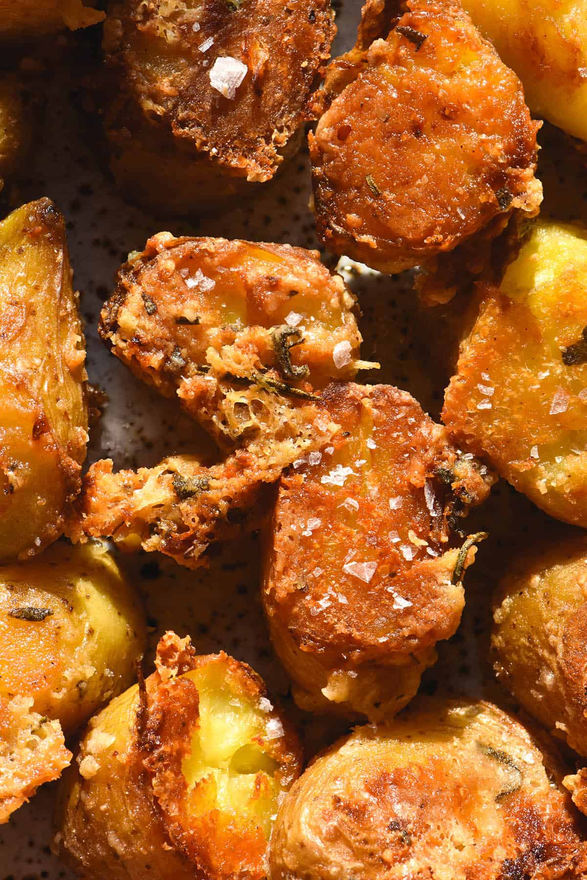 An aerial macro image of crispy roasted kipfler potatoes with a parmesan and herb crust. The potatoes are golden brown and sprinkled with sea salt flakes