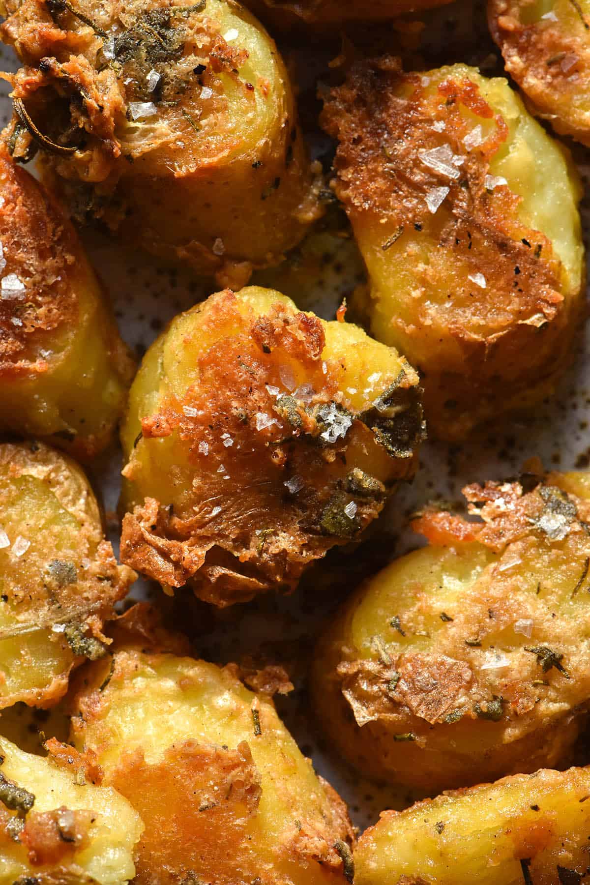 An aerial macro view of roasted kipfler potatoes with a parmesan herb crust on a white speckled ceramic plate. The potatoes are golden brown and topped with a parmesan herb crust. 