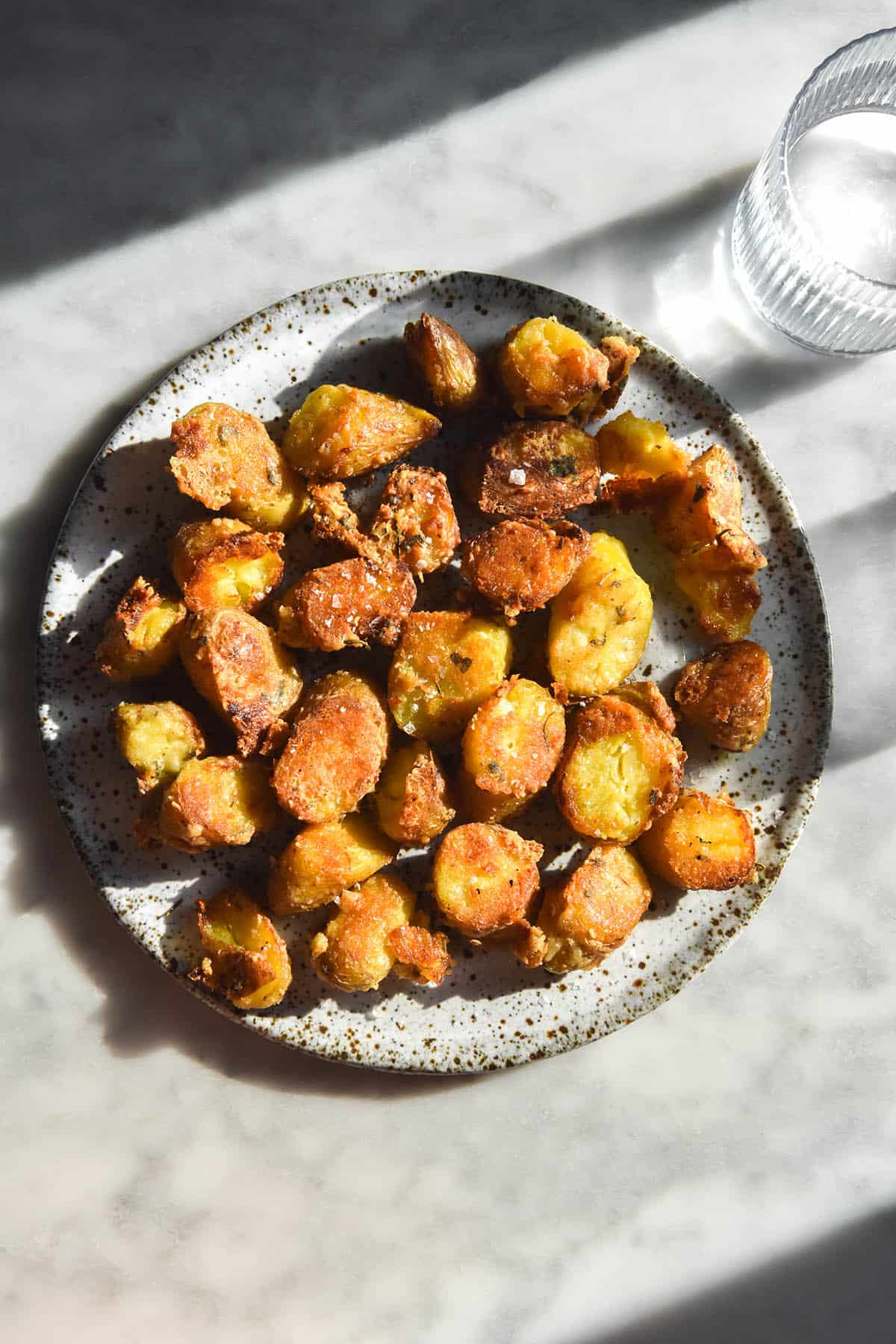 An aerial image of a white speckled ceramic plate topped with roasted kipfler potatoes with a parmesan herb crust. The plate sits atop a white marble table bathed in contrasting sunlight and shadow, and two water glasses sit to the right of the potatoes