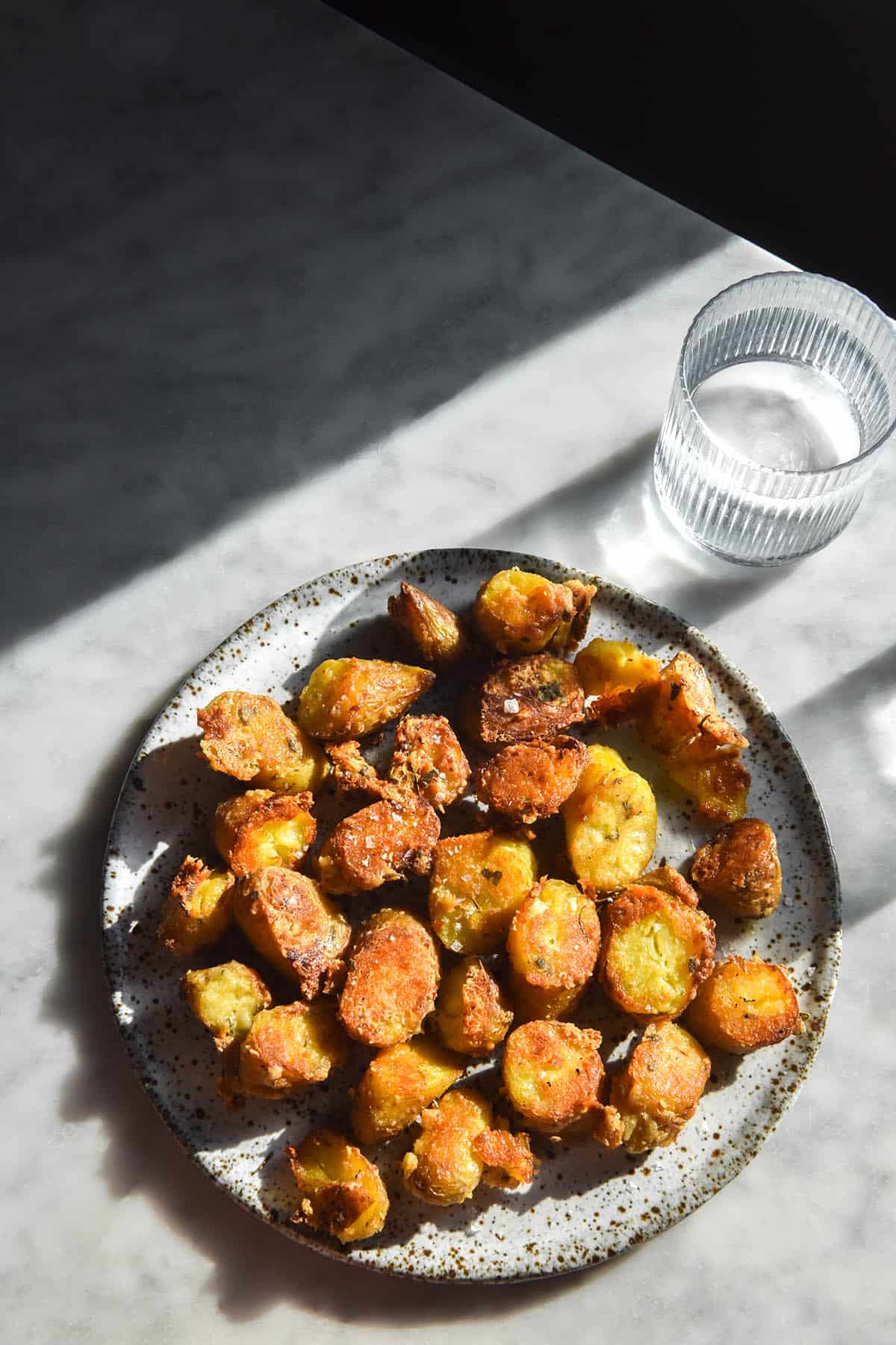 An aerial image of crispy roasted kipfler potatoes with a parmesan herb crust atop a white speckled ceramic plate. The plate sits on the bottom half of a white marble table, which is bathed in contrasting sunlight. The top third of the image is out of the sun, creating an angular contrasting pattern on the table. 