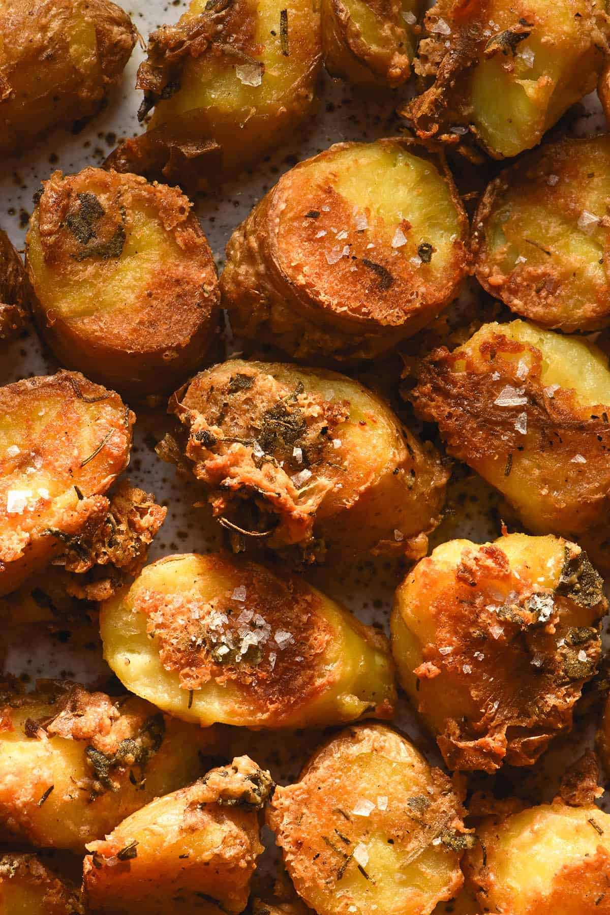 An aerial macro image of a plate of roasted kipfler potatoes with a herb and parmesan crust. The potatoes are golden brown and topped with sea salt flakes