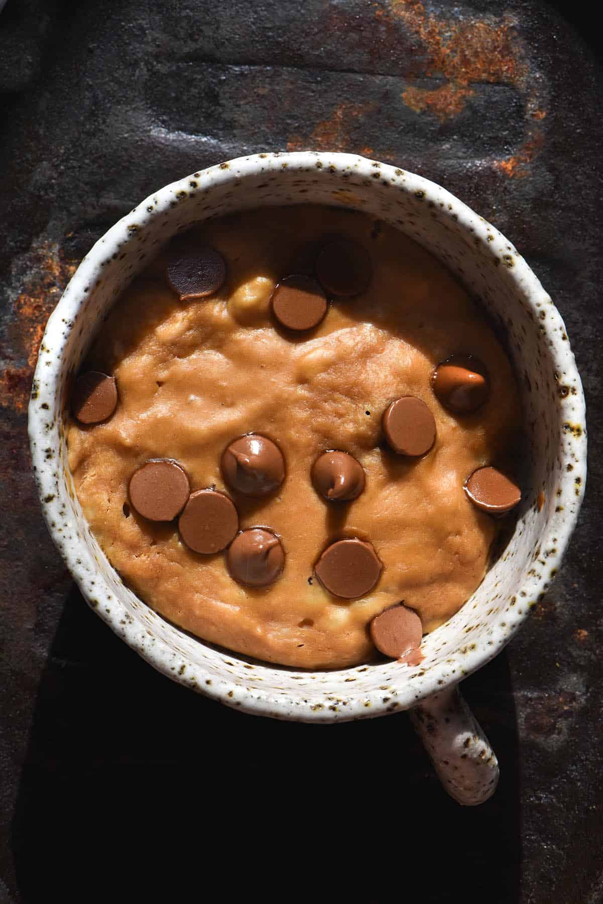 An aerial image of a white speckled ceramic mug filled with a vegan protein mug cake topped with chocolate chips. The mug sits atop a lightly rusted steel platter. 