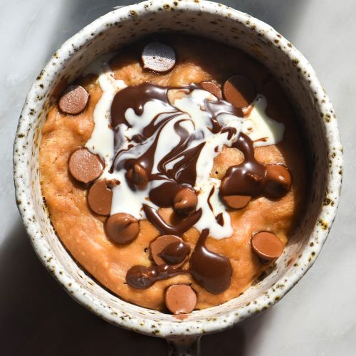 An aerial view of a vegan protein mug cake in a white speckled ceramic mug atop a white marble table. The mug cake is topped with chocolate chips, vegan Nutella and a swirl of coconut cream