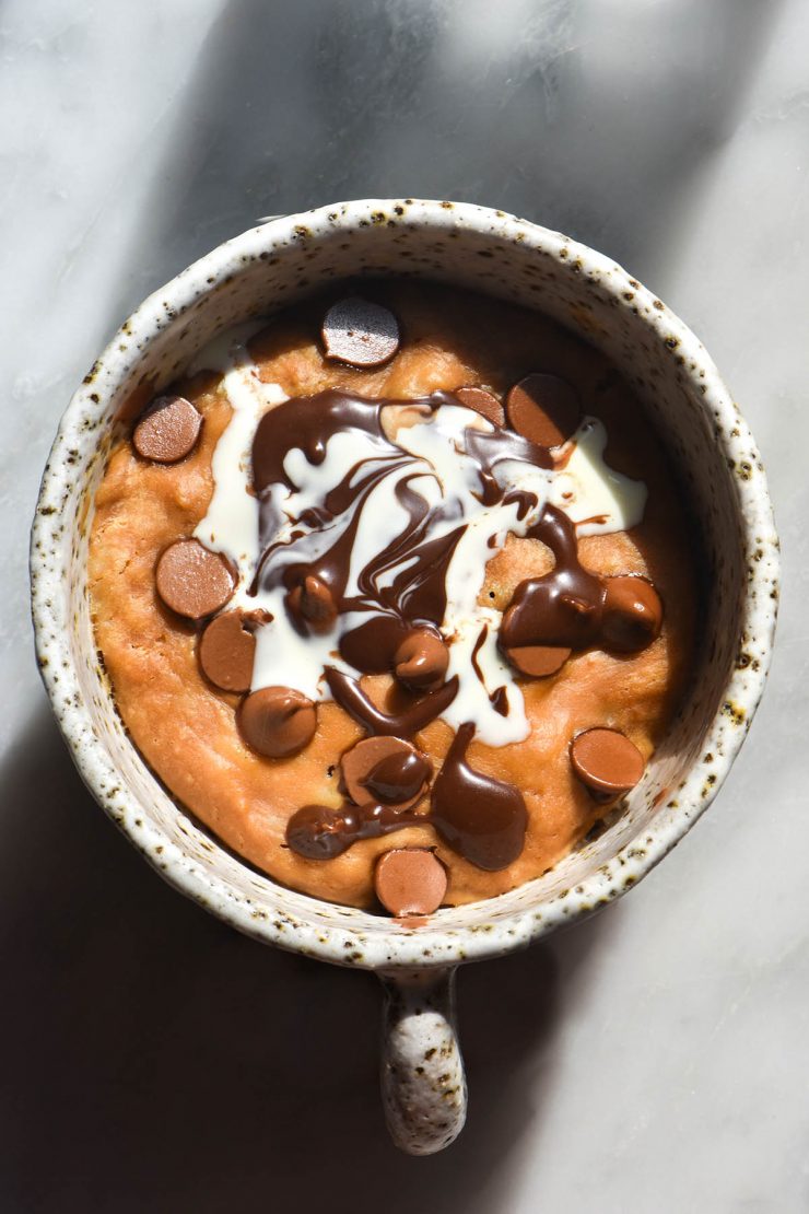 An aerial view of a vegan protein mug cake in a white speckled ceramic mug atop a white marble table. The mug cake is topped with chocolate chips, vegan Nutella and a swirl of coconut cream