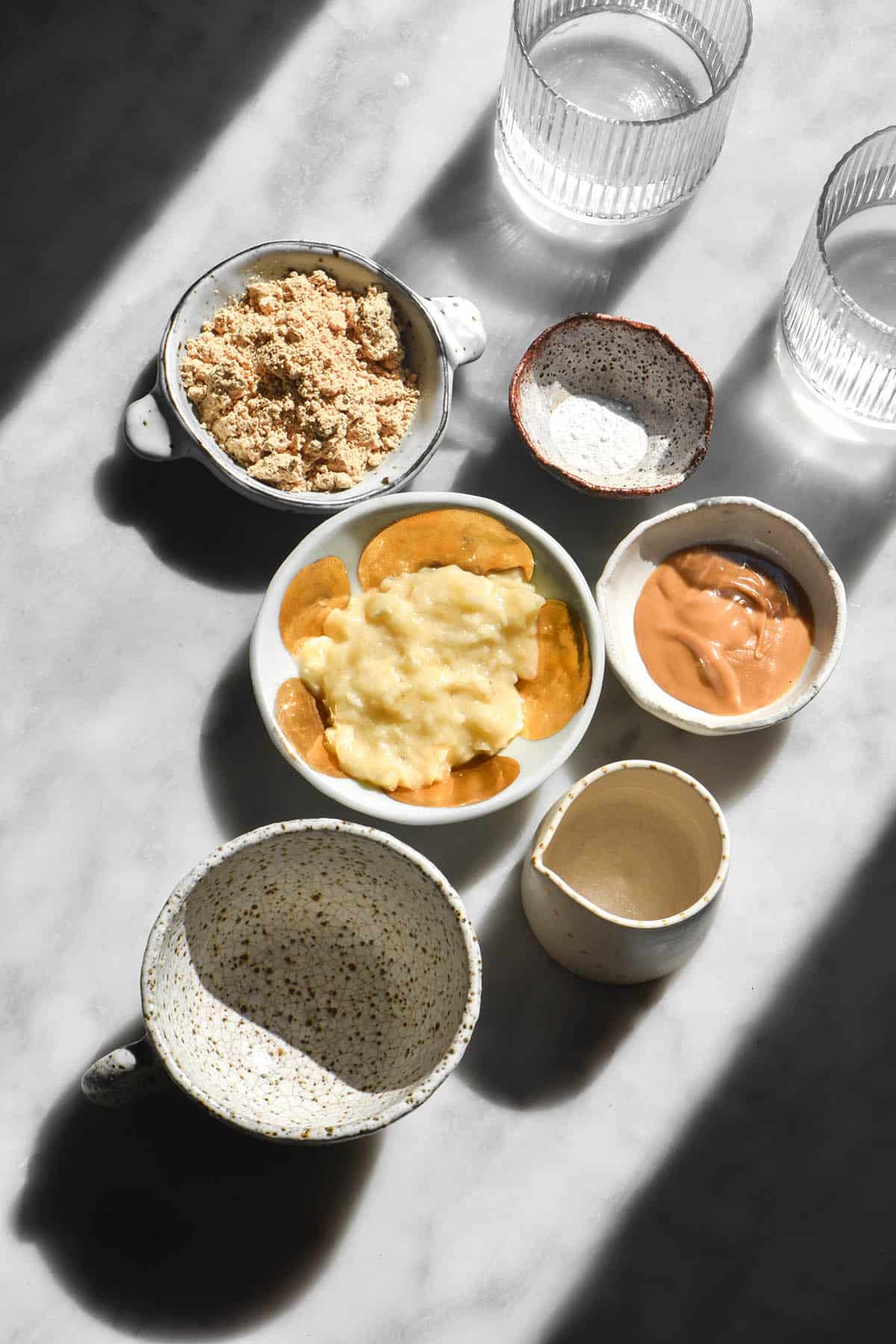 An aerial image of the ingredients needed to make a vegan protein mug cake. The ingredients are all in small white ceramic bowls, and are set on a white marble table in contrasting sunlight. 