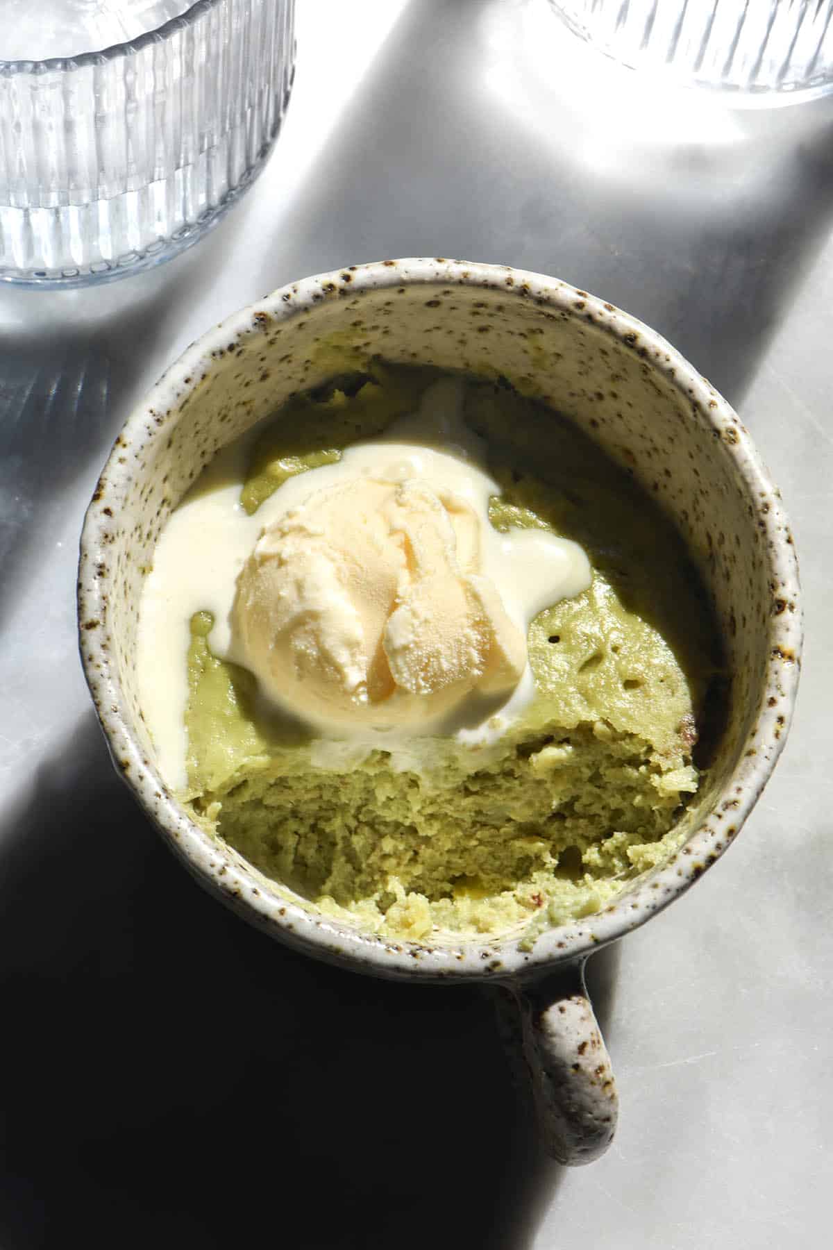 A close up aerial image of a vegan protein mug cake made with pumpkin seed protein. The mug cake is a matcha green and topped with melting vanilla ice cream. It sits atop a sunlit white marble table with glasses of water in the background 