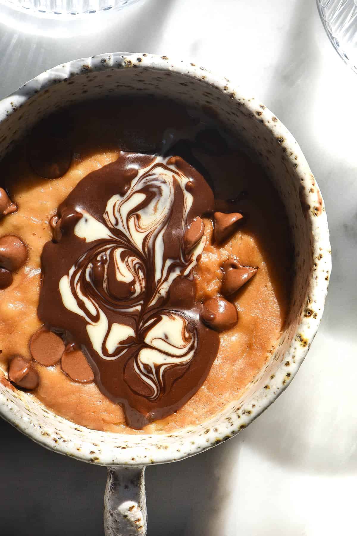 An aerial close up view of a vegan protein mug cake topped with chocolate chips and swirled vegan Nutella and coconut cream. The mug cake sits atop a white marble sunlit table. 