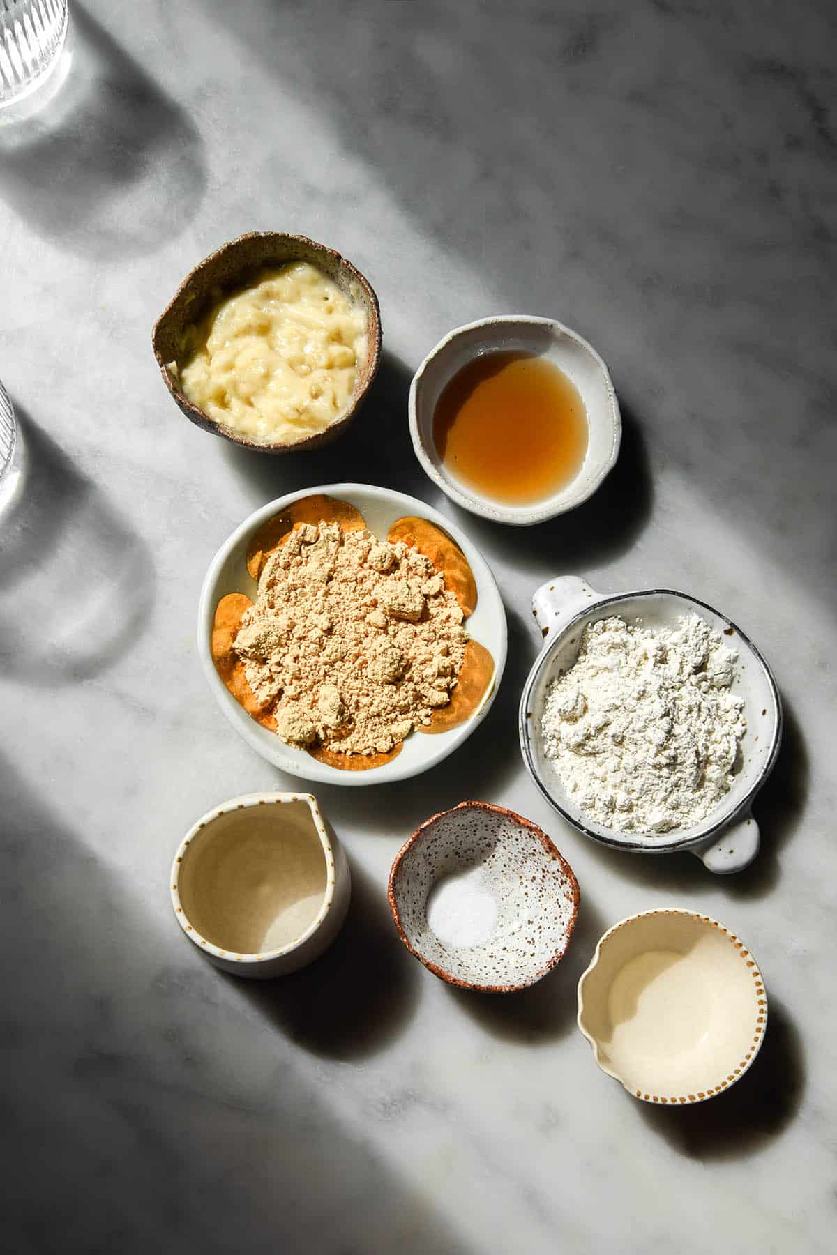 An aerial view of small pinch bowls filled with the ingredients needed to make vegan protein pancakes atop a white marble table in contrasting sunlight