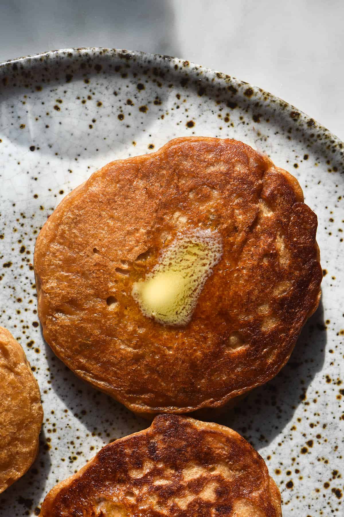 A close up aerial macro photo of three vegan protein pancakes on a white speckled ceramic plate. The central pancake is topped with a small pat of butter that is melting into the golden pancake