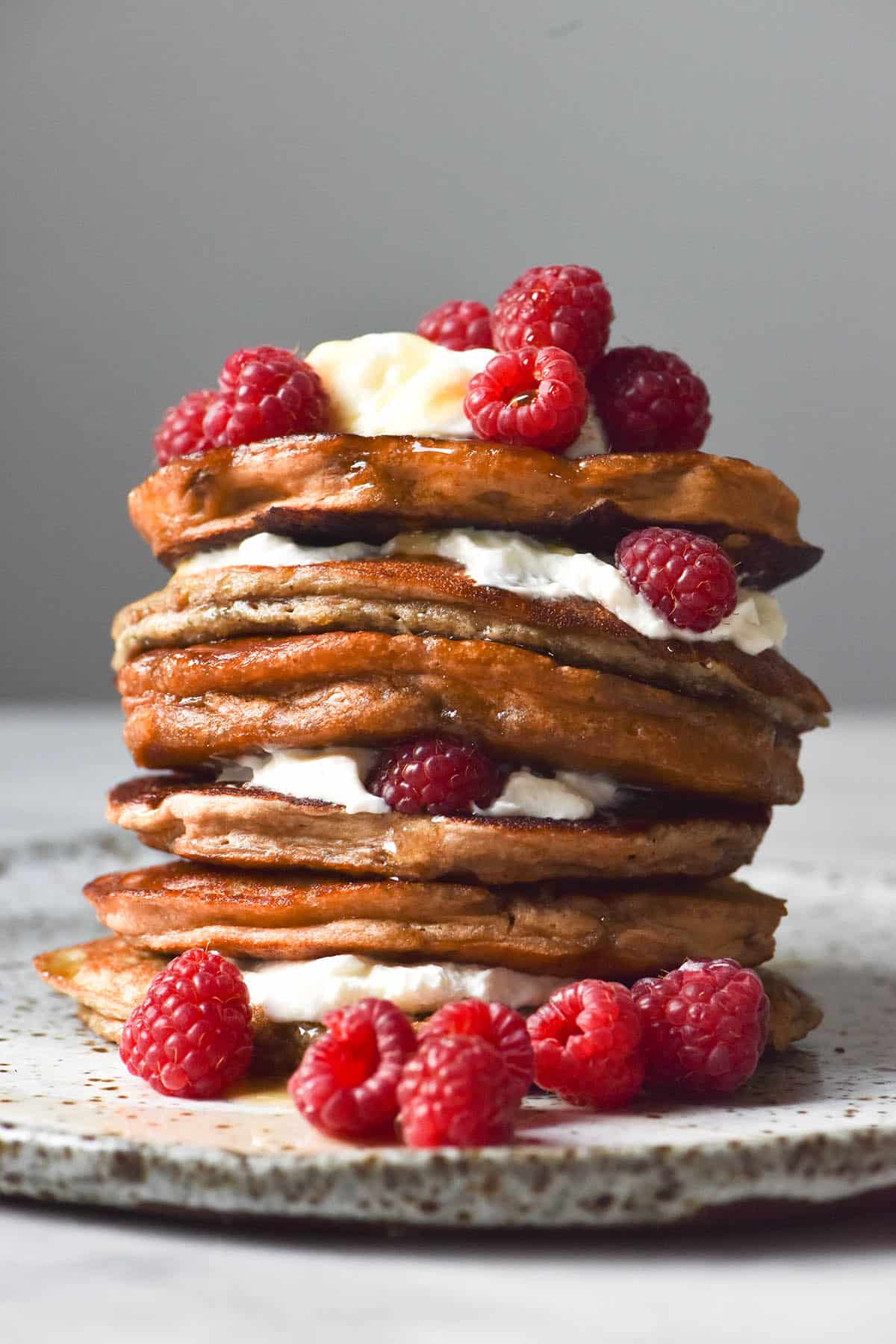 A side on view of a stack of vegan protein pancakes stuffed with yoghurt and raspberries and drizzled with maple syrup. The pancakes sit on a white speckled ceramic plate against a white backdrop