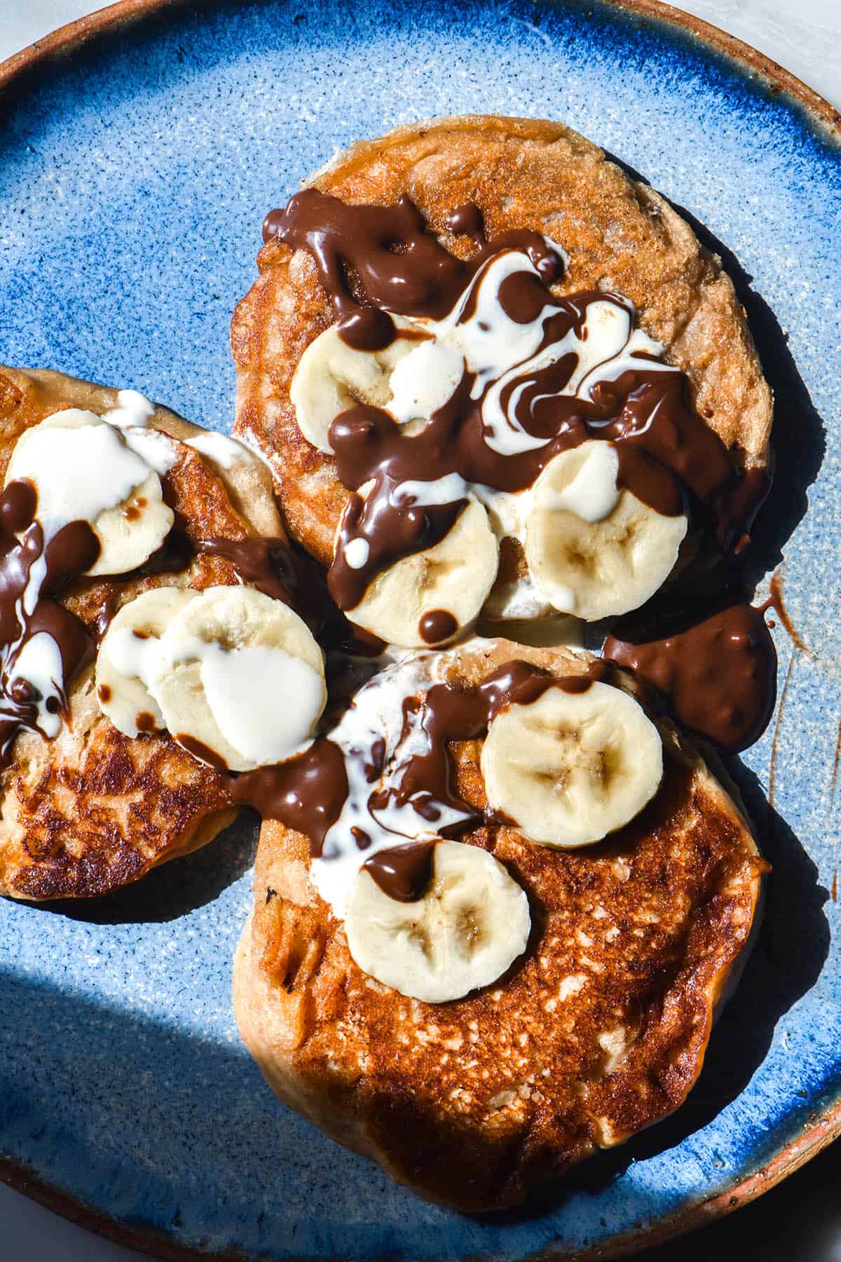 An aerial view of three gluten free vegan protein pancakes on a bright blue plate in bright sunlight. The pancakes are topped with vegan nutella, coconut cream and extra slices of banana