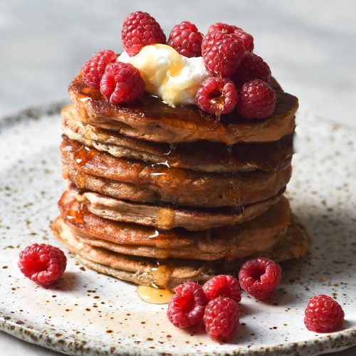 A side on image of a stack of vegan protein pancakes on a white speckled ceramic plate against a white backdrop. The pancakes are topped with yoghurt, raspberries and a drizzle of maple syrup.