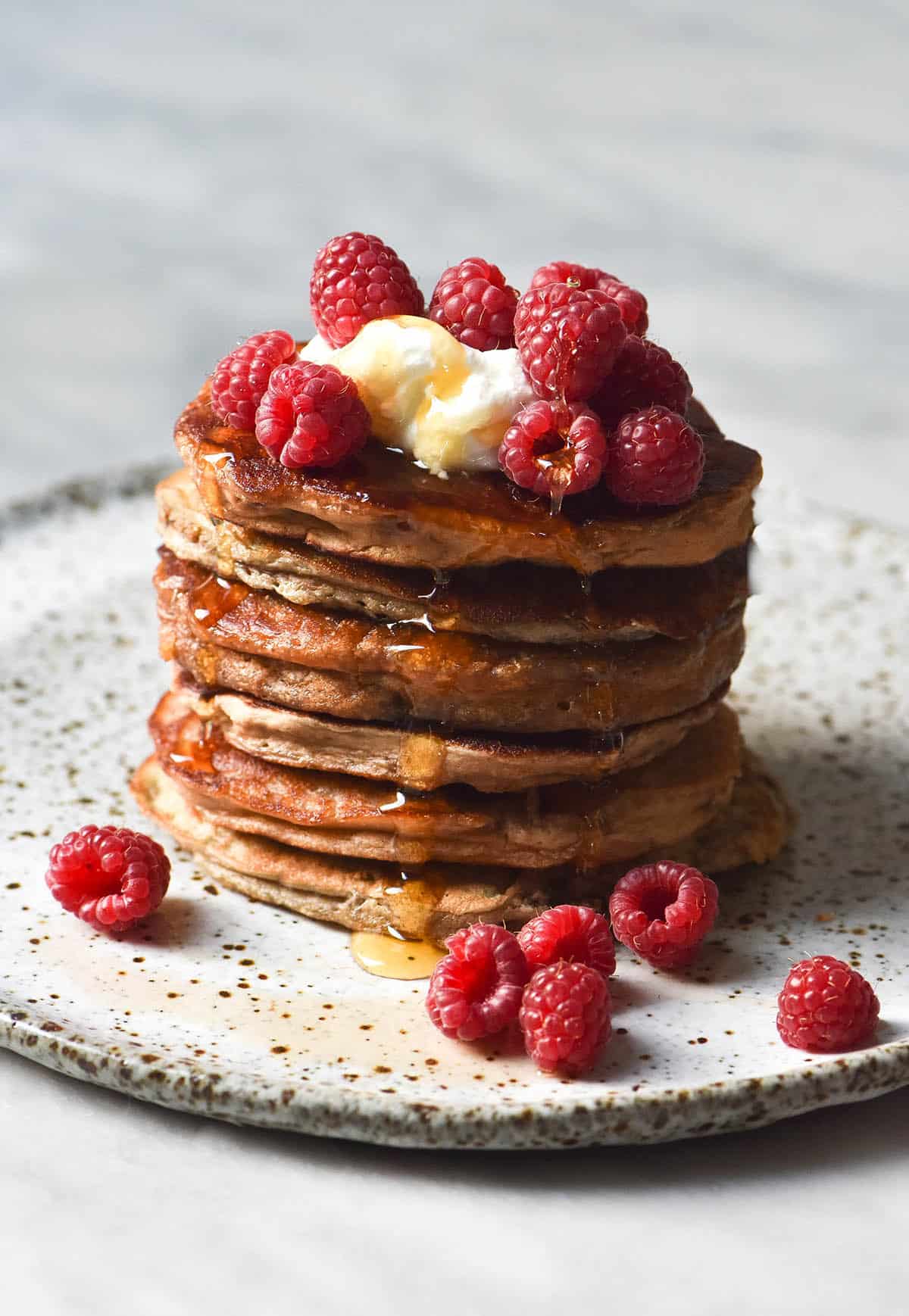 A side on image of a stack of vegan protein pancakes on a white speckled ceramic plate against a white backdrop. The pancakes are topped with yoghurt, raspberries and a drizzle of maple syrup. 