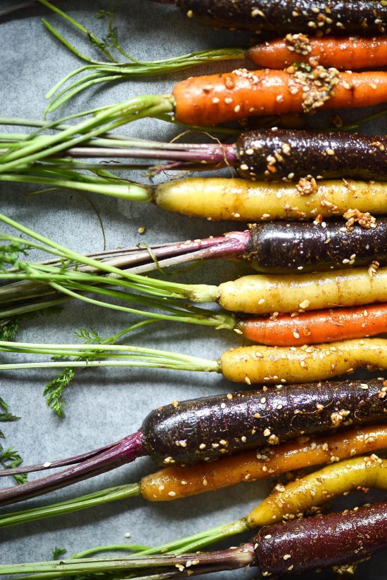 An aerial macro image of heirloom, multicoloured carrots covered in oil and dukkah, lined up on a baking tray, their greens to the left