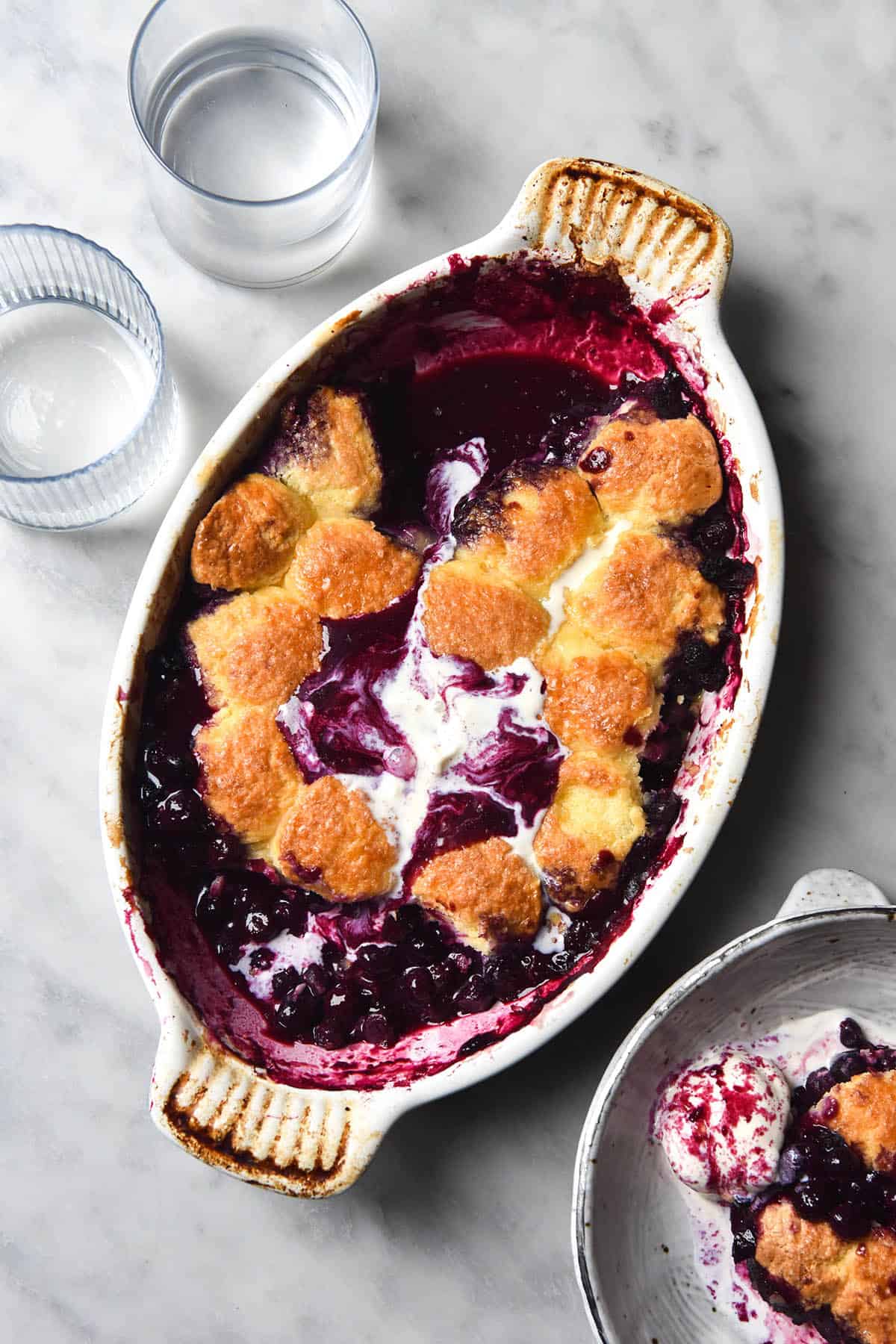 An aerial image of a gluten free blueberry cobbler in an oval white ceramic baking dish. The cobbler pieces are golden brown and melted vanilla ice cream swirls together with blueberry juice to create a marbled pattern on top of the cobbler