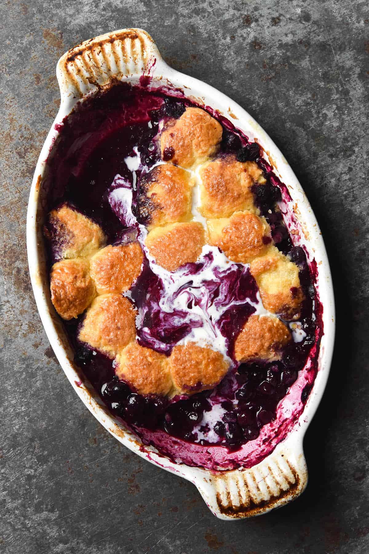 An aerial image of a gluten free blueberry cobbler in an oval white ceramic baking dish on a blue grey mottled steel backdrop. The cobbler pieces are golden brown and melted vanilla ice cream swirls together with blueberry juice to create a marbled pattern on top of the cobbler