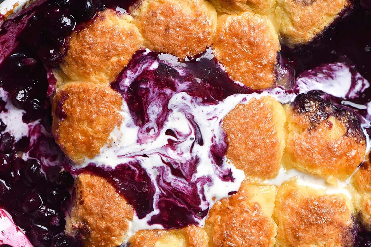 A macro close up image of a gluten free blueberry cobbler topped with melted vanilla ice cream. The cobbler is golden brown and the vanilla swirled with deep purple blueberry juice creates a marbled pattern on the top of the cobbler 