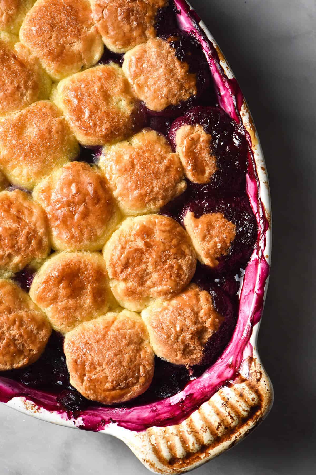 A close up aerial image of a gluten free blueberry cobbler in a white ceramic baking dish atop a white marble table. The cobbler topping is golden brown and the deep purple blueberry juices have create a bright purple pattern on the edges of the baking dish