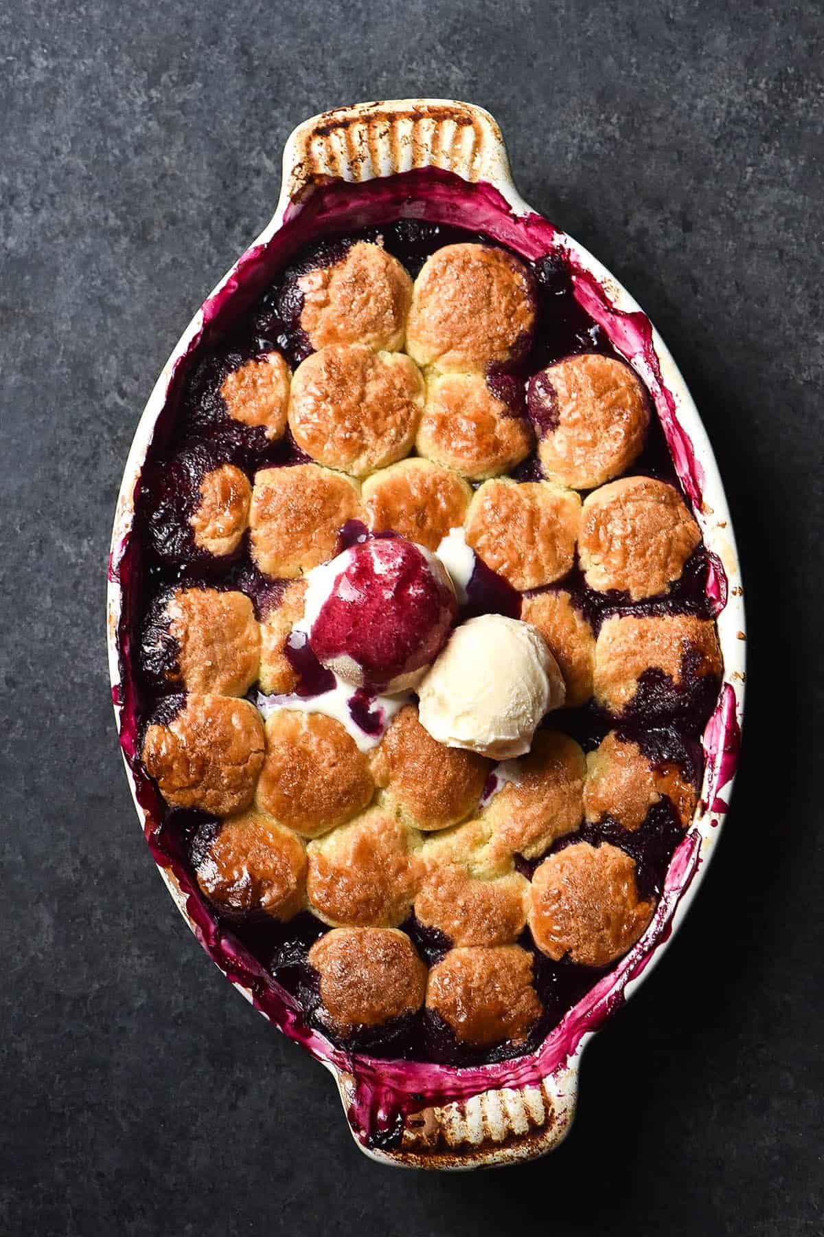 An aerial image of a gluten free blueberry cobbler on a dark blue backdrop. The cobbler is topped with two scoops of vanilla ice cream, and one has been covered in deep purple blueberry juices