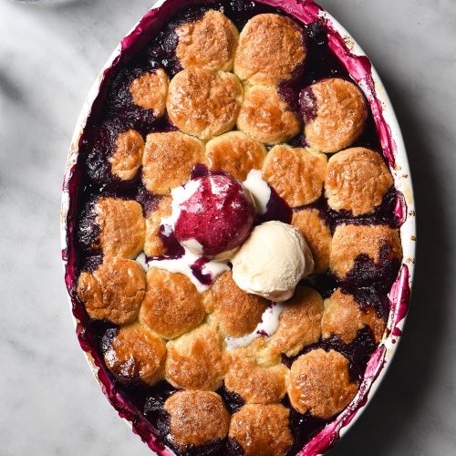 An aerial image of a gluten free blueberry cobbler on a dark blue backdrop. The cobbler is topped with two scoops of vanilla ice cream, and one has been covered in deep purple blueberry juices