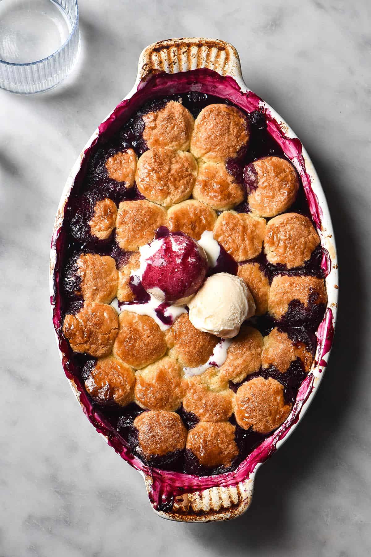 An aerial image of a gluten free blueberry cobbler on a white marble table. The cobbler is topped with two scoops of vanilla ice cream, and one has been covered in deep purple blueberry juices