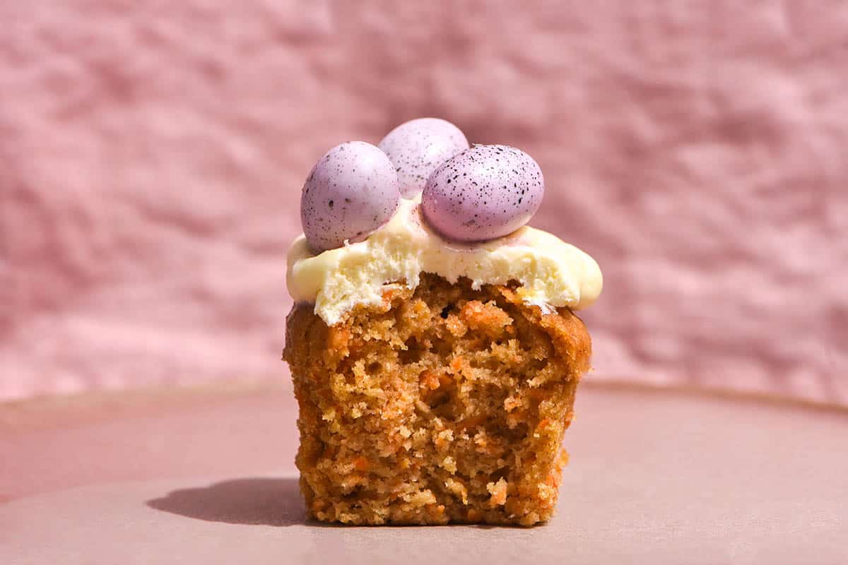 A brightly lit side on view of a gluten free carrot cake muffin topped with cream cheese icing and pastel speckled eggs. The muffin sits atop a pale pink ceramic plate against a pale pink textured linen backdrop