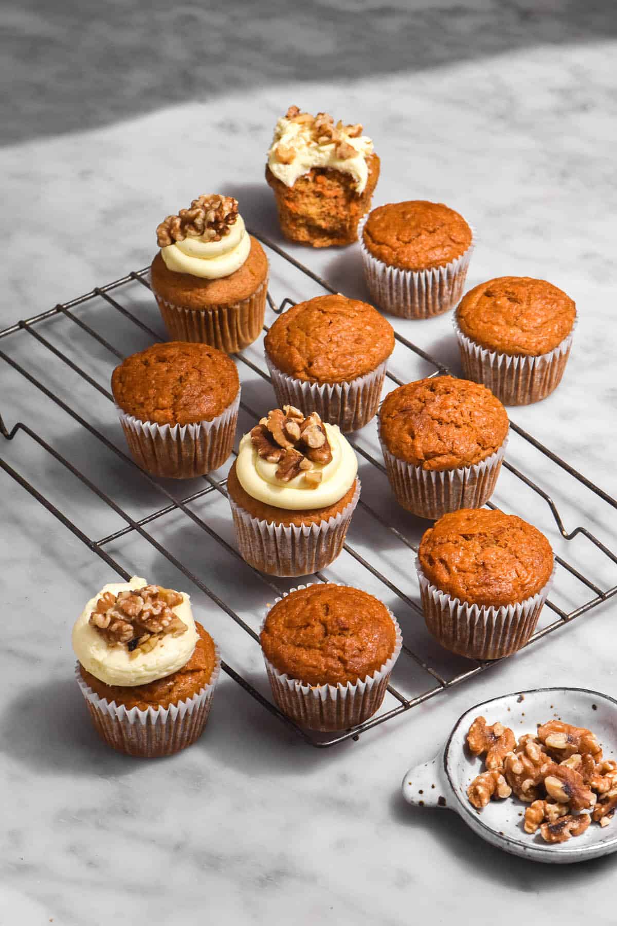 A brightly lit image of gluten free carrot cake muffins on a cooling rack atop a white marble table. Some of the muffins are topped with cream cheese icing and chopped walnuts. 