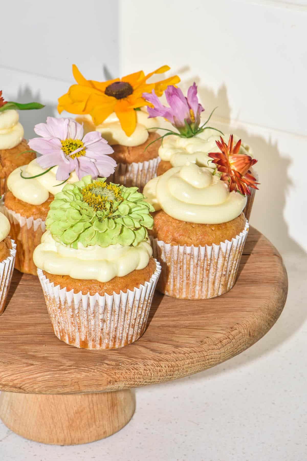 A brightly lit side on flash image of gluten free carrot cupcakes on a wooden cake stand against a white stone kitchen backdrop. The muffins are topped with piped cream cheese and a different coloured spring flower for each cupcake.