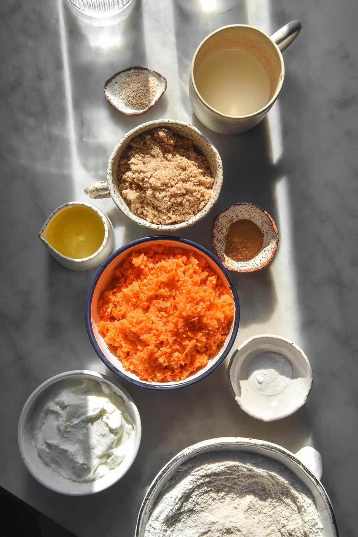 An aerial image of the ingredients used to make gluten free carrot muffins. The muffins sit in small white bowls atop a white marble table in contrasting sunlight. 