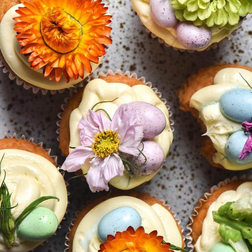 An aerial image of gluten free carrot muffins topped with cream cheese icing, flowers and pastel mini Easter eggs