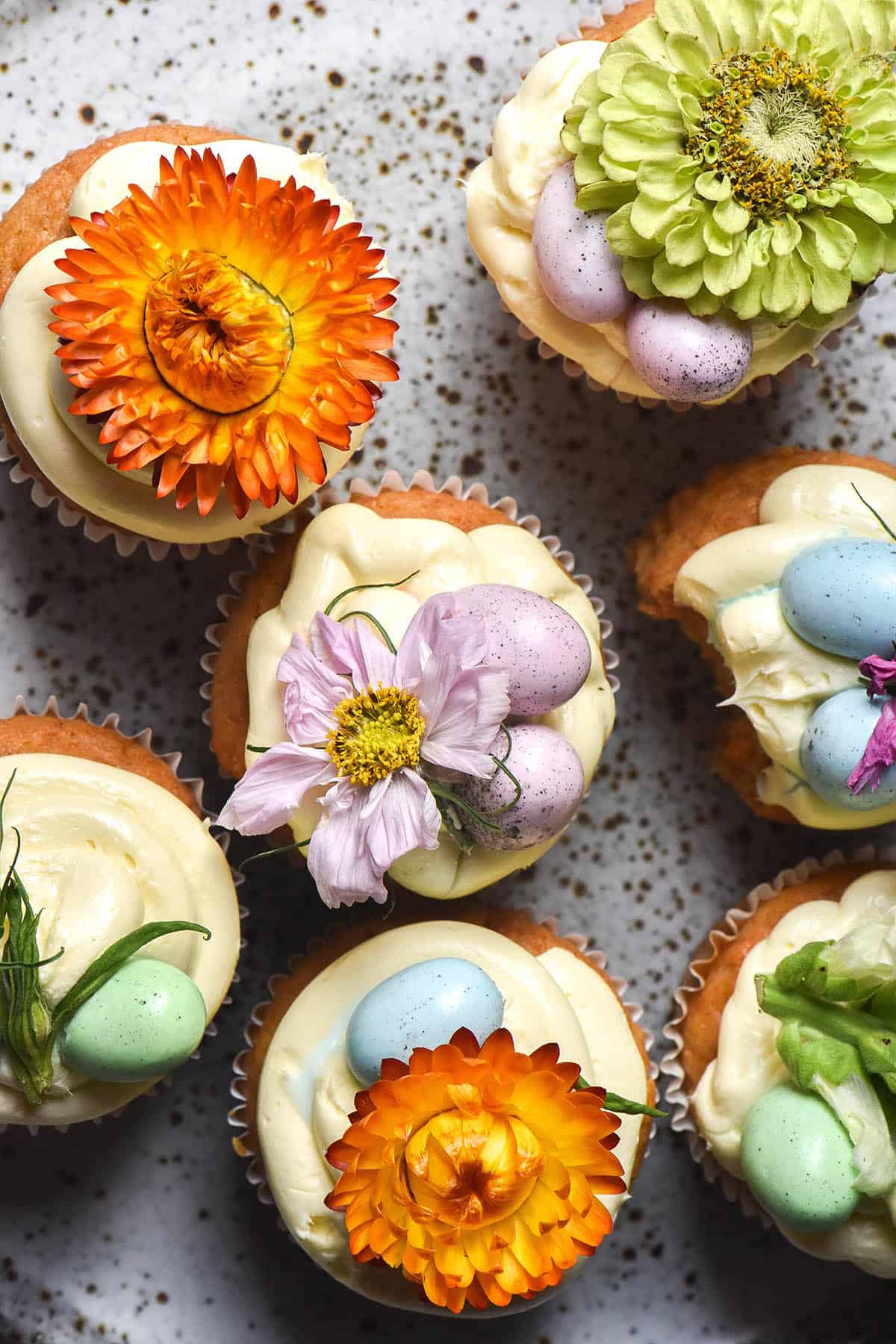 An aerial image of gluten free carrot muffins topped with cream cheese icing, flowers and mini pastel Easter eggs. They sit on a white speckled ceramic plate. 