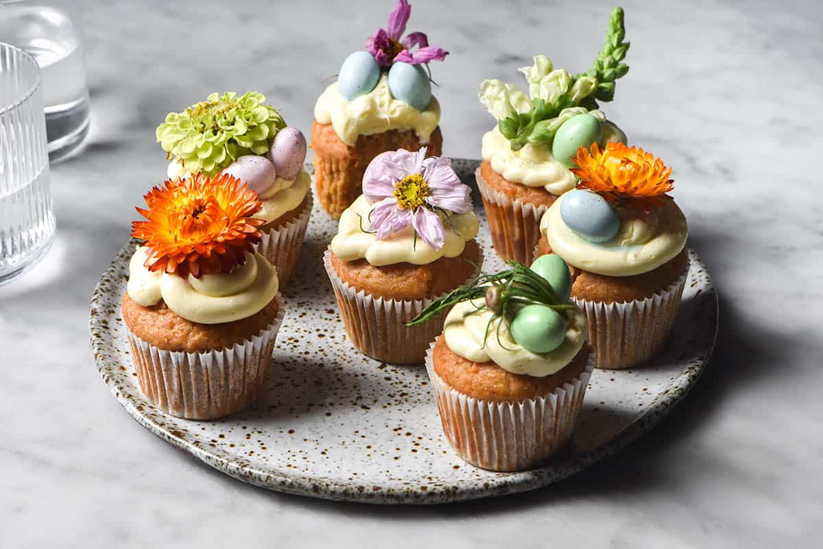A side on image of a white speckled ceramic plate topped with gluten free carrot muffins. The muffins are topped with cream cheese icing, flowers and pastel mini Easter eggs. 