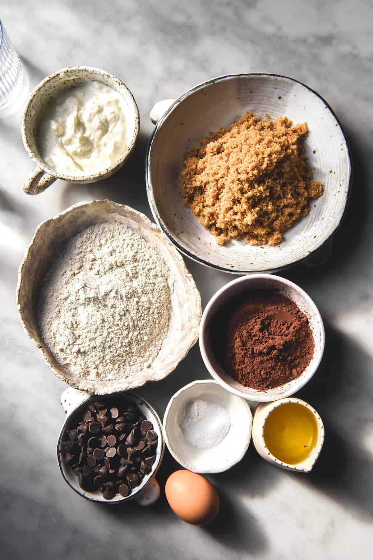 An aerial image of the ingredients used to make gluten free chocolate muffins. They are grouped together in small white bowls on a white marble table. 