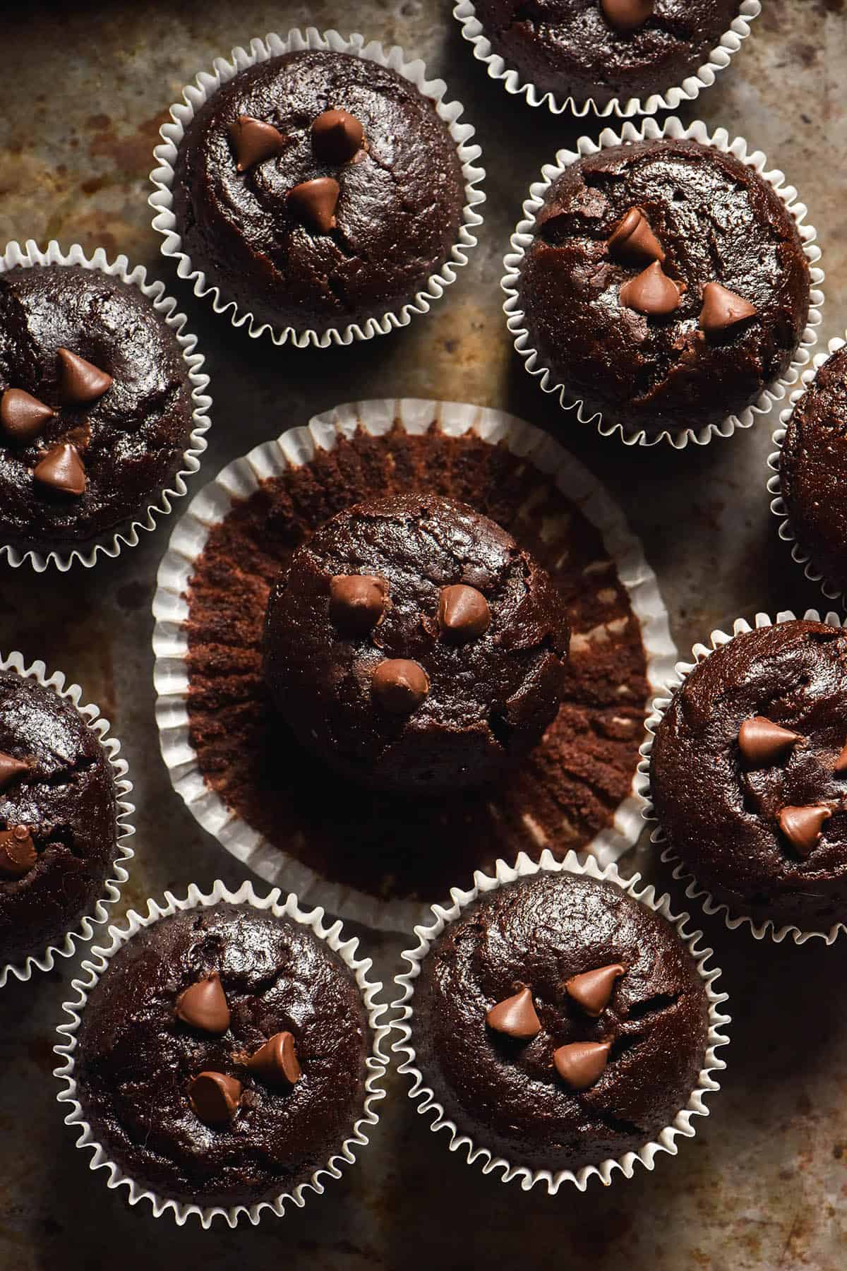 An aerial image of gluten free chocolate muffins sitting on a dark steel backdrop. The central muffin has had the liner peeled away from the top, creating a flower like circle from above. 