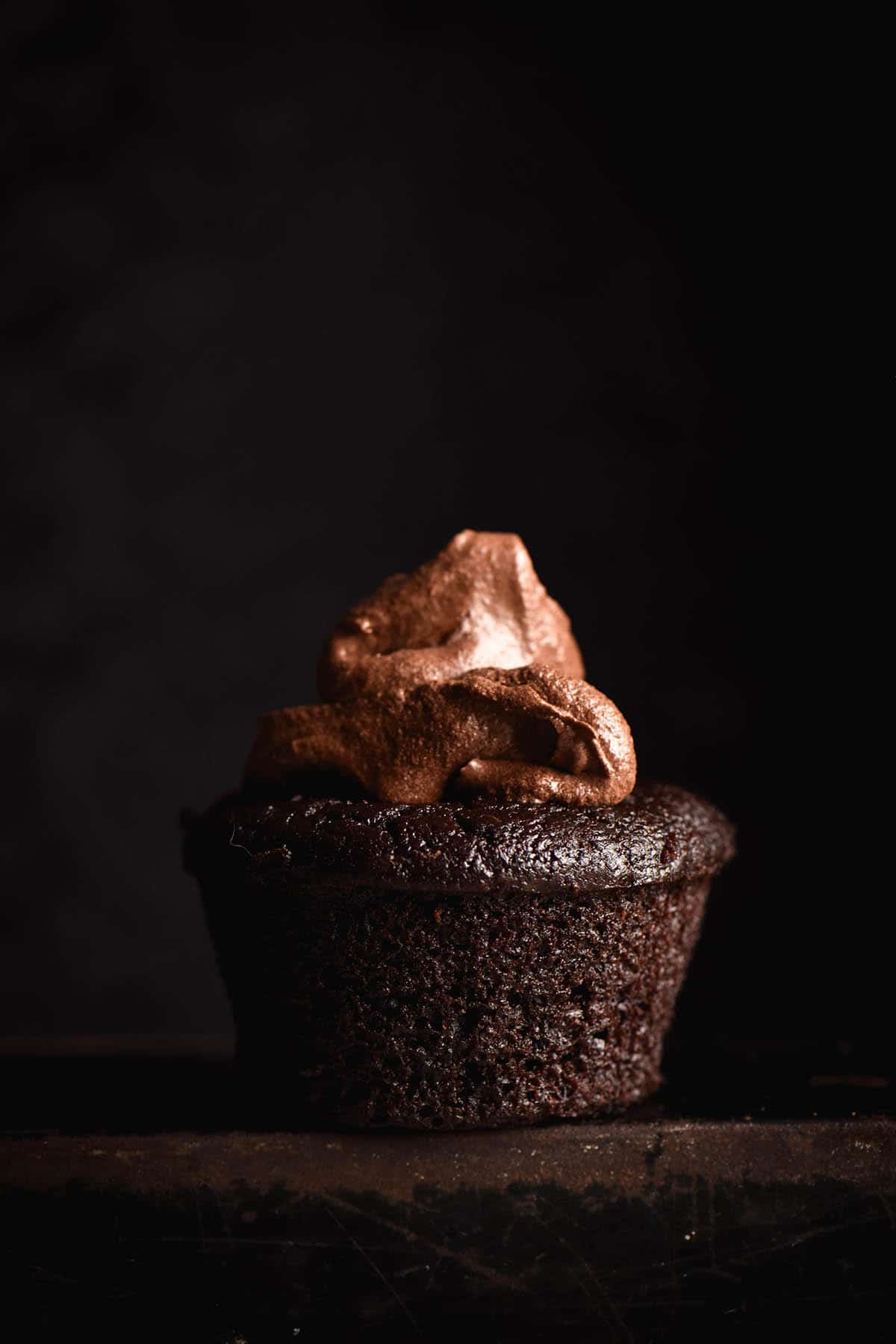 A side on dark and moody image of a gluten free chocolate muffin against a black backdrop. The muffin is topped with a casual dollop of chocolate buttercream.