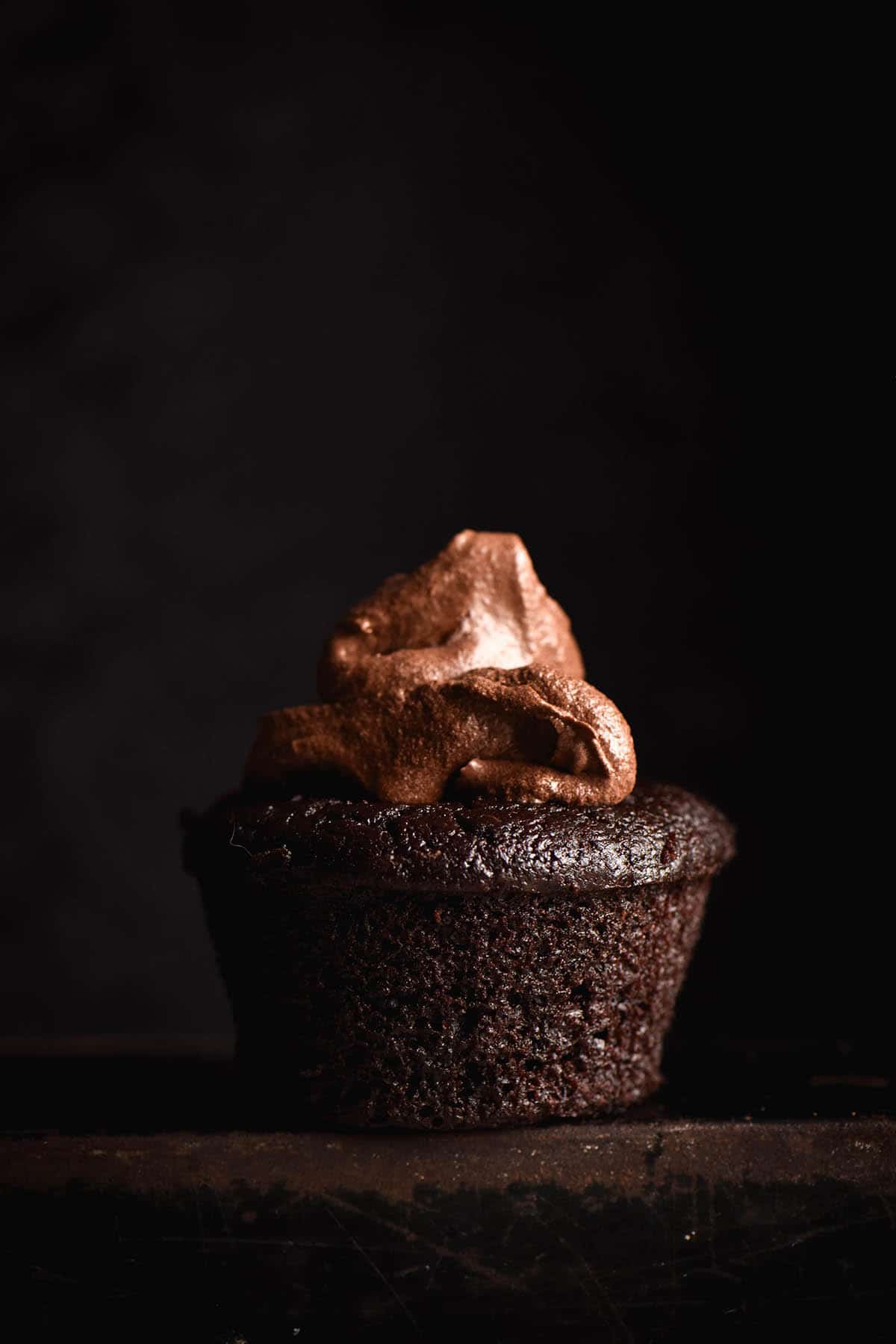 A dark and moody side on image of a gluten free chocolate muffin topped with chocolate buttercream icing