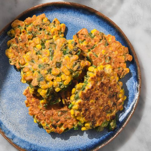 An aerial image of a bright blue ceramic plate topped with gluten free corn fritters. The plate sits on a white marble table with a water glass to the top left of the plate.