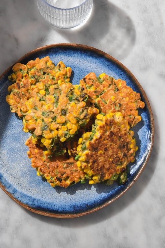 An aerial image of a bright blue ceramic plate topped with gluten free corn fritters. The plate sits on a white marble table with a water glass to the top left of the plate.