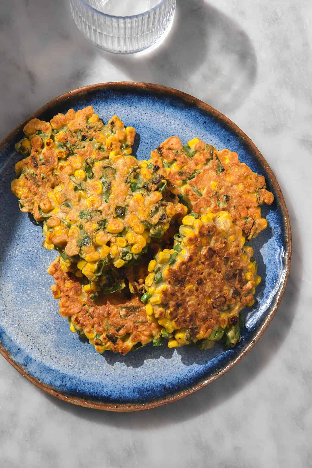 An aerial image of a bright blue ceramic plate topped with gluten free corn fritters. The plate sits on a white marble table with a water glass to the top left of the plate. 