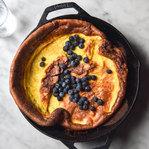An aerial view of a gluten free Dutch baby topped with blueberries in a black skillet on a white marble table.