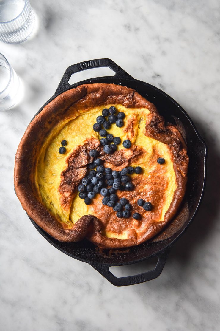 An aerial view of a gluten free Dutch baby topped with blueberries in a black skillet on a white marble table.