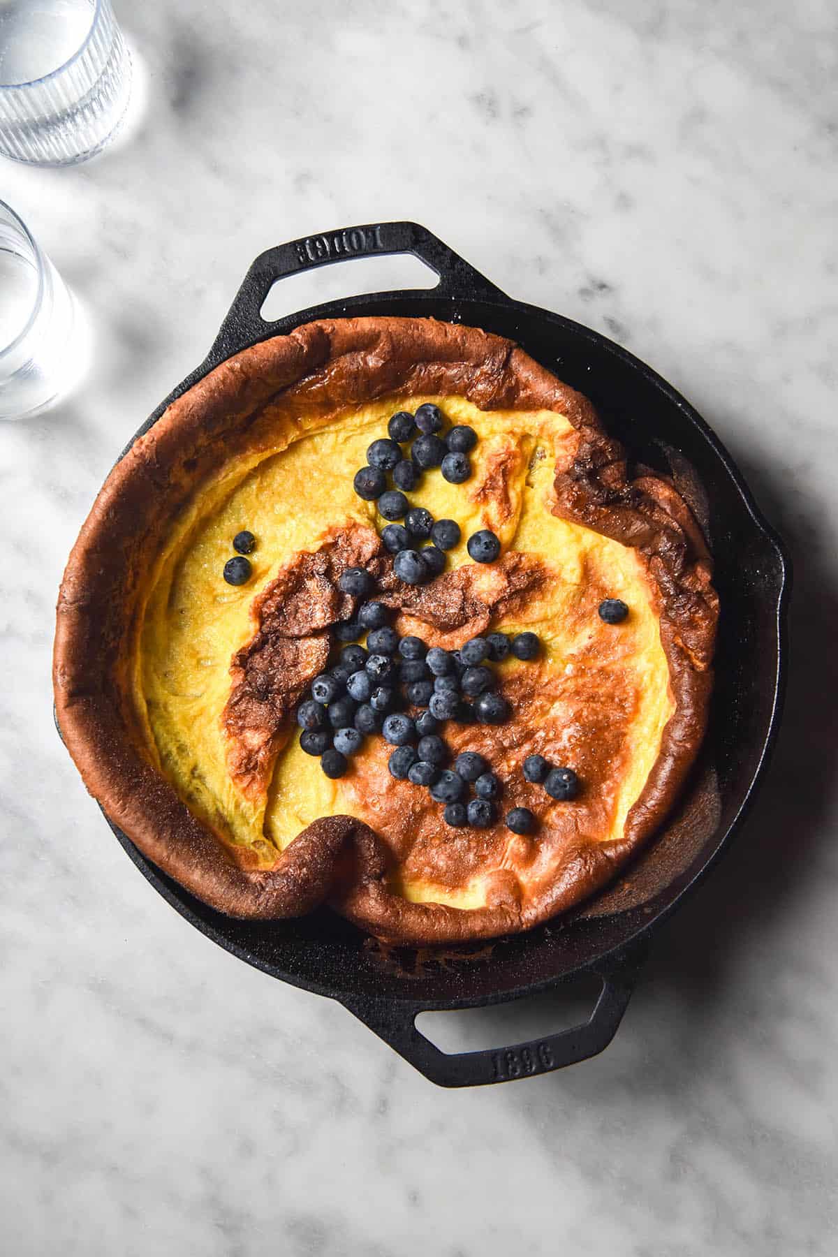 An aerial view of a gluten free Dutch baby topped with blueberries in a black skillet on a white marble table. 