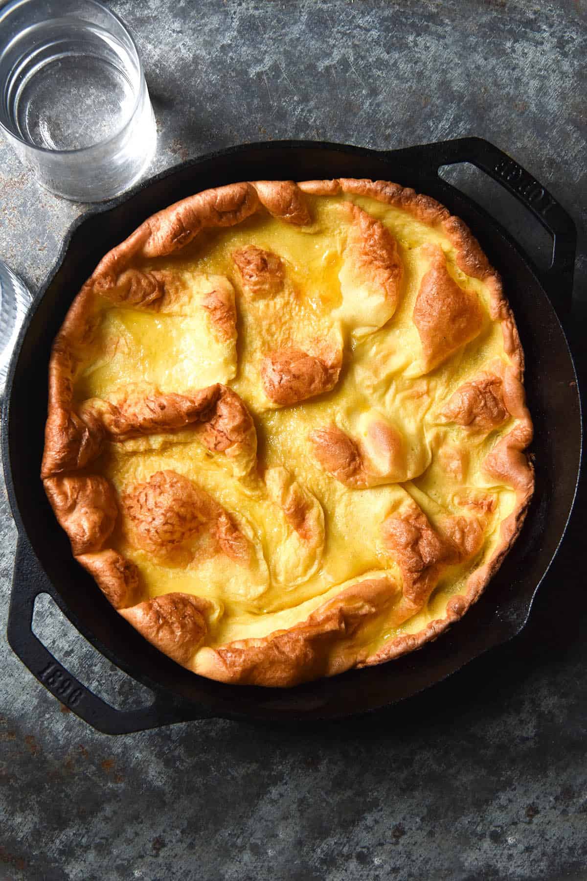 An aerial image of a gluten free dutch baby in a black skillet atop a dark steel backdrop. Two glasses of water sit to the left of the skiller