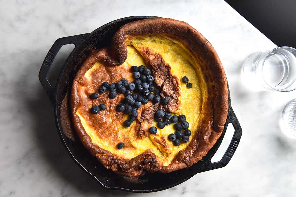 An aerial view of a gluten free Dutch baby topped with blueberries in a black skillet on a white marble table. 
