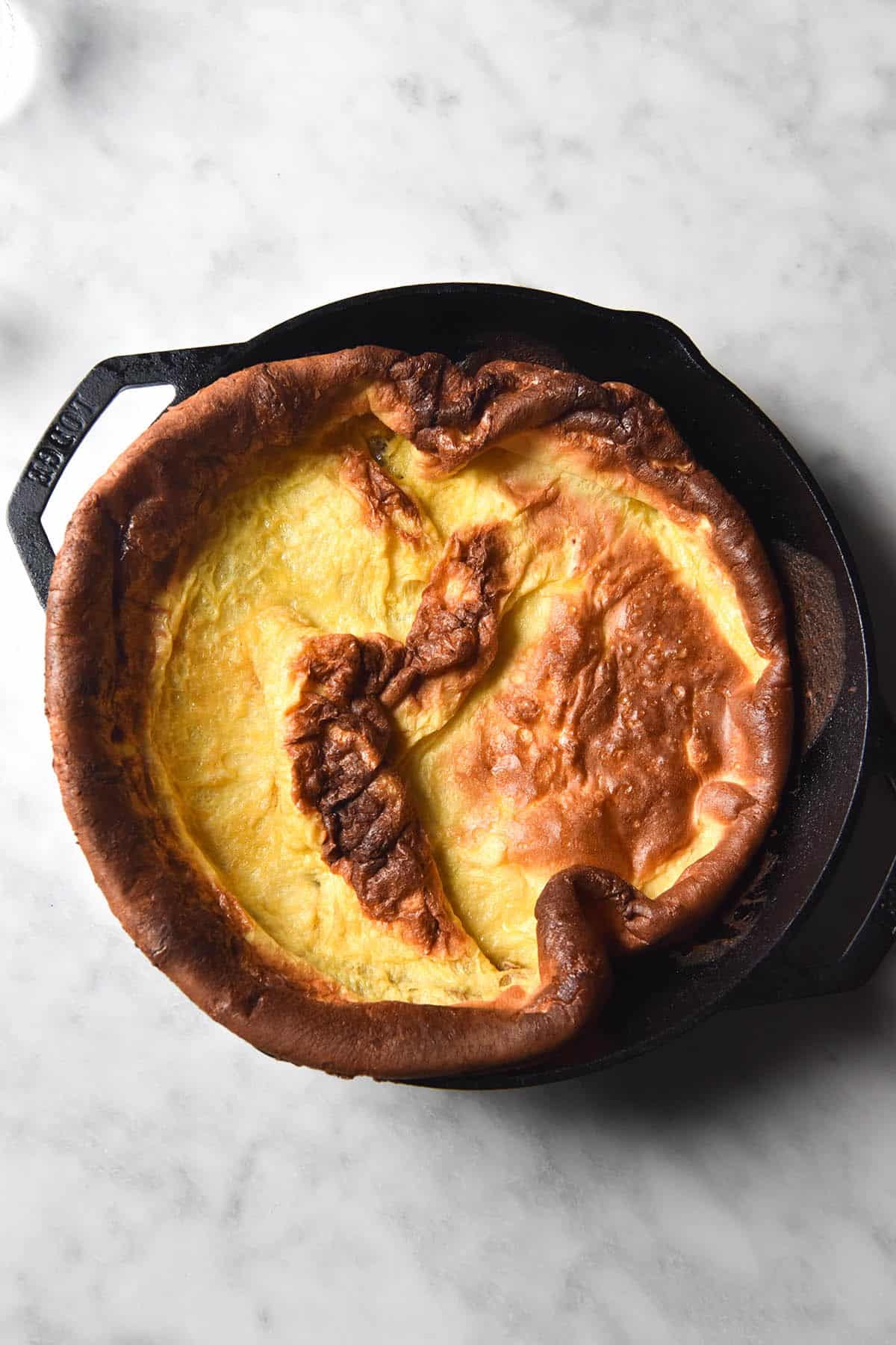 An aerial image of a gluten free Dutch baby in a black skillet atop a white marble table. 