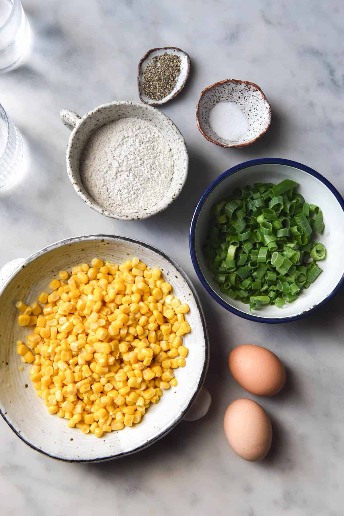 An aerial image of the ingredients used for gluten free corn fritters. The ingredients are arranged in small white bowls of varying sizes on a white marble table. Two glasses of water sit to the top left of the image. 