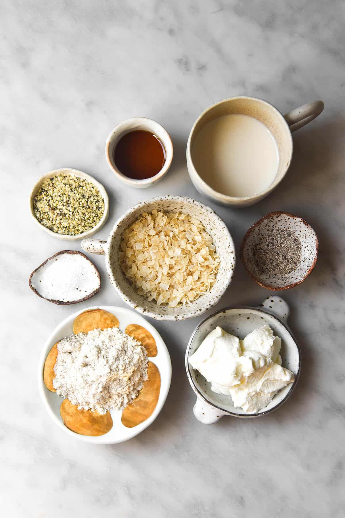 An aerial image of the ingredients for gluten free overnight oats grouped in small white ceramic bowls on a white marble table