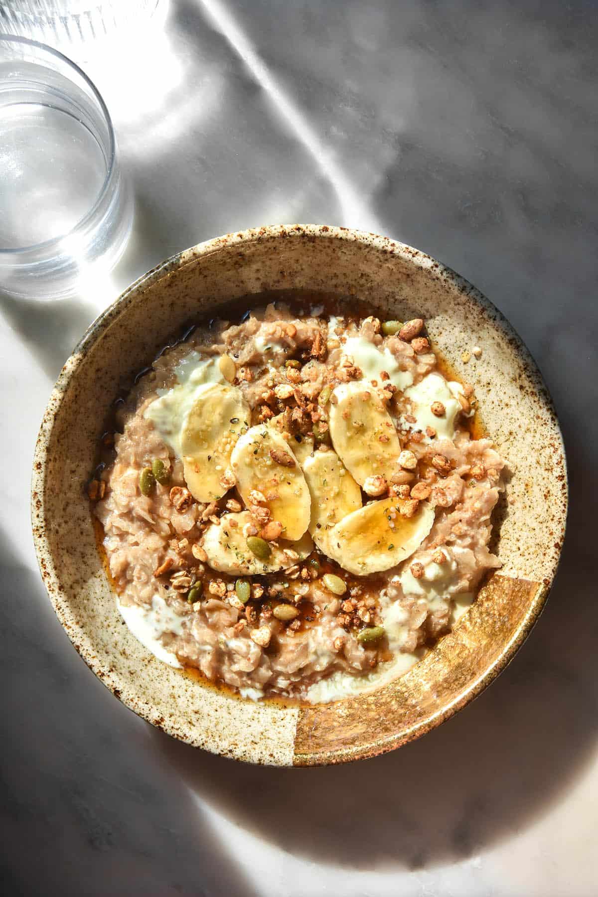 An aerial sunlit image of a bowl of gluten free overnight oats topped with bananas, granola and some yoghurt. The bowl sits in contrasting sunlight on a white marble table. Two glasses of water sit to the top left of the image and cast a shadow across the bowl.