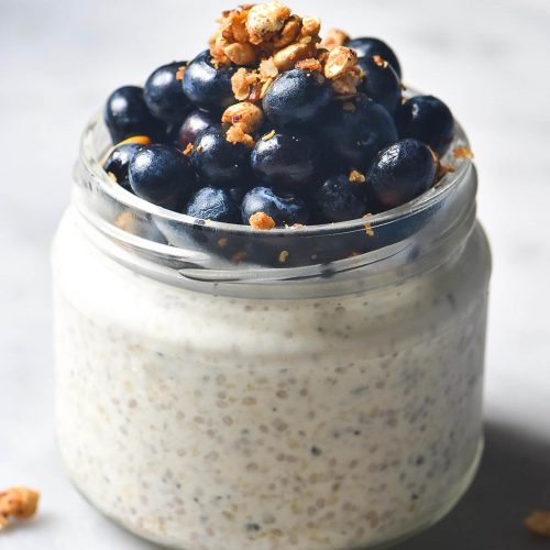 A close up side on image of a round jar filled with gluten free overnight oats and topped with blueberries and a sprinkle of granola. The jar sits on a white marble table and a sprinkle of granola surrounds the jar.