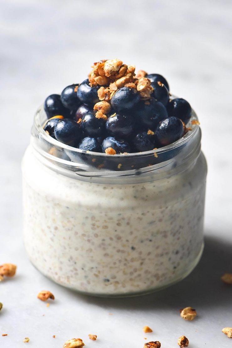 A close up side on image of a round jar filled with gluten free overnight oats and topped with blueberries and a sprinkle of granola. The jar sits on a white marble table and a sprinkle of granola surrounds the jar.